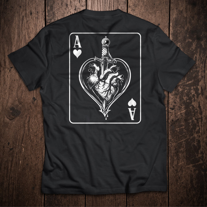 Black t-shirt flat lay on wooden surface with Ace of Hearts playing card design featuring anatomical heart pierced by sword illustration