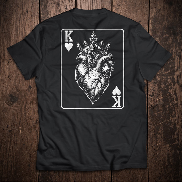 Black t-shirt flat lay on wooden surface with King of Hearts playing card design featuring crowned anatomical heart illustration