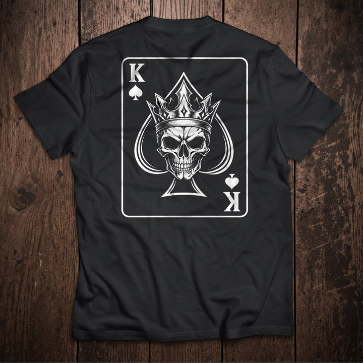 Black t-shirt flat lay on wooden surface with King of Spades playing card design featuring crowned skull illustration