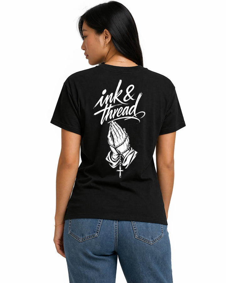 Female model showing back of black t-shirt with Ink and Thread script calligraphy and praying hands rosary illustration on white background