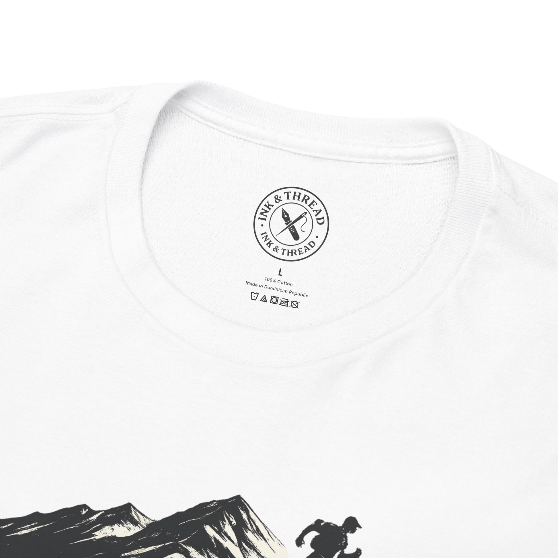 Close-up of Ink and Thread UK inner neck label on white t-shirt featuring mountain and hiker graphic, showing brand stamp size and care instructions